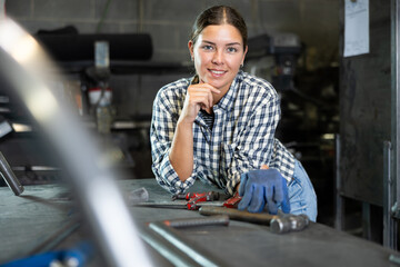Confident young female worker in casual checkered shirt and gloves, standing in metal workshop with slight smile, ready for work..