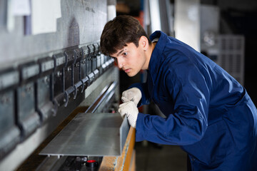 Focused young male worker in blue work coat and gloves operating press brake in industrial...