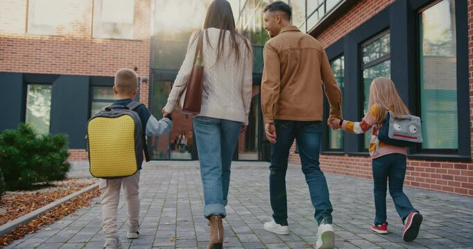 Camera view from behind of Caucasian family walking together towards entrance to school building. Looking after children. Holding hands with small kids. Children carrying backpacks.