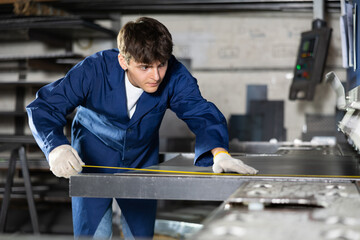Guy working at metallurgical plant carefully measures sheet of iron using a tape measure