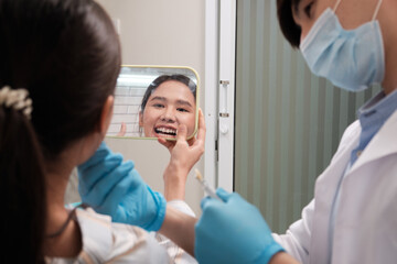 Asian male dentist explains color shades of tooth implant whitening to young female patient, look at mirror in dental clinic, and do professional mouth hygiene healthcare work in a doctor's hospital.