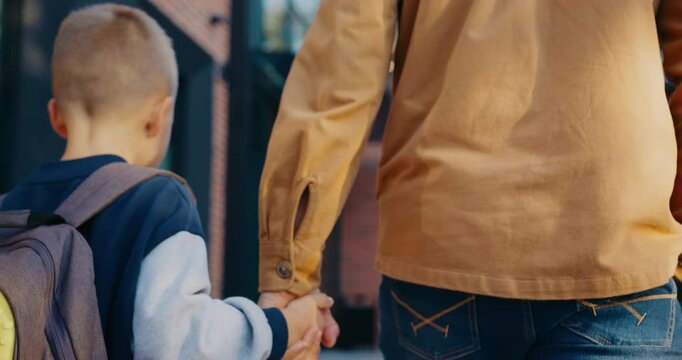 View from behind of small Caucasian boy walking together with his caring dad. Protective father dropping off small boy at school. Holding his hand. Carrying backpack with books. Modern school.