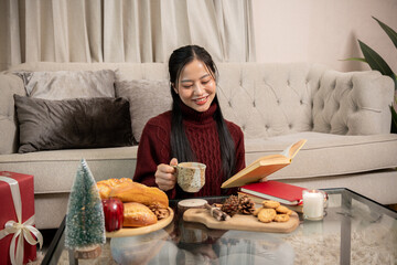 Asian woman in red sweater sitting reading a book and sipping a warm drink in living room during...