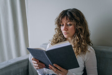 Young woman sitting comfortably on a gray sofa, engrossed in reading a book, with a soft, natural...