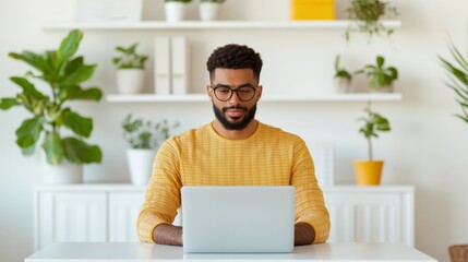 A man in a yellow sweater works on a laptop in a bright room adorned with plants and shelves.