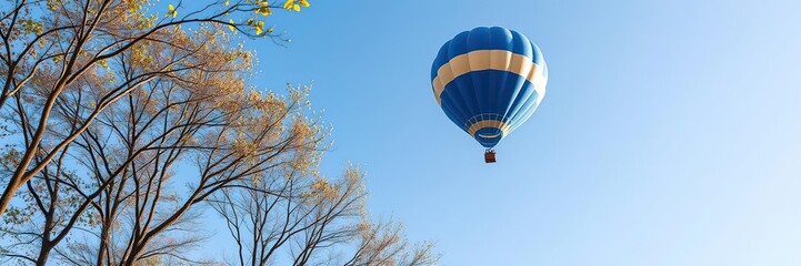 Blue and gold flying balloon soaring above the trees on a clear blue day with a few leaves, green landscape, blue and gold flying balloon