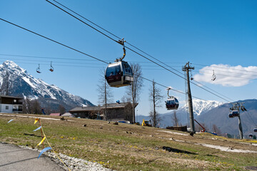 Cable cars glide above mountainous terrain on a clear day, offering views of snowy peaks and green pathways near a ski resort - Rosa Khutor, Russia April 15, 2023