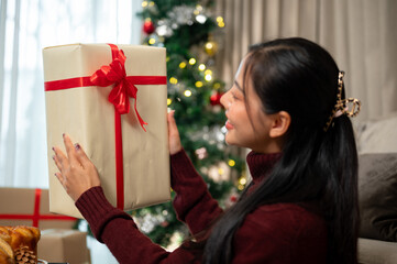 A happy Asian woman sits in the living room, admiring her Christmas present with a joyful smile.