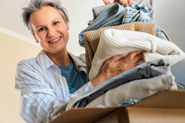 Portrait of smiling gray haired mature woman pack clothes pile to cardboard box for donation