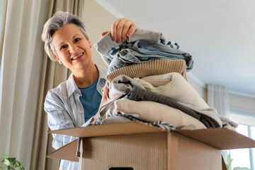 Portrait of smiling gray haired mature woman pack clothes pile to cardboard box for donation