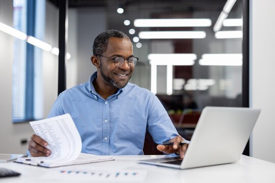 Mature African American businessman working on laptop in office, reviewing documents and displaying focus and professionalism. Concept of work, business analysis, modern technology.