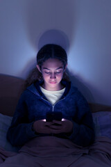 Sad and tired teenager girl using a smartphone lying in bed late at night, playing games, watching...