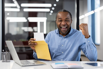 Mature African American businessman celebrates success at office desk with laptop and open envelope, displaying joy and accomplishment. Image conveys excitement, achievement, and career progress.