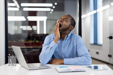 Mature African American businessman yawning at office desk with open laptop. Depicts workplace...