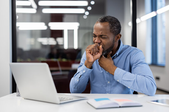 Mature African American businessman sitting at desk in modern office, coughing into hand. Concerned expression indicates health issues affecting work.