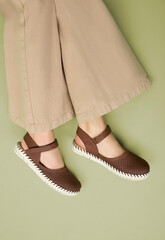 Close-up of a young woman modeling brown woven Mary Jane flats with adjustable straps, white stitched soles, beige wide-leg trousers, a muted green background for a cozy autumn-inspired fashion look.