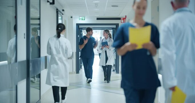 Diverse Medical Personnel, Including a Caucasian Male ICU Doctor in Blue Scrubs and Female General Practitioner in a White Coat, Walking in Busy Hallway while Having a Casual Healthcare Conversation