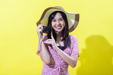 Indonesian Asian woman in a straw hat and pink floral dress, smiling and holding a camera with both hands against a yellow background. Bright, even lighting.