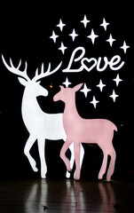 Two illuminated deer silhouettes, one white and one pink, stand side by side on a dark background with the word Love and a scattering of stars above them.