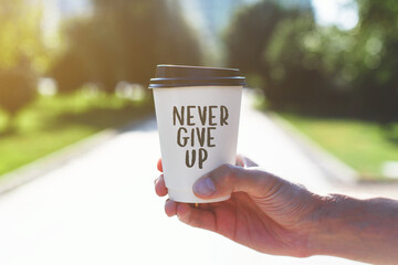 A hand holding a takeaway coffee cup with the motivational quote "Never Give Up" printed on it. Bright sunlight, blurred green park background, positive and inspiring atmosphere.