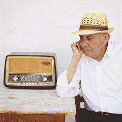 An older gentleman wears a straw hat while sitting at a wooden table. He looks thoughtfully at a vintage radio, enjoying the nostalgic sound on a bright day.