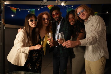Multiethnic friends toasting at rooftop party with colorful string lights