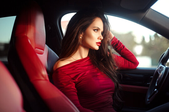 Beautiful Smiling woman driving car in red, attractive girl sitting in automobile, indoors summer portrait. Sexy brunette model with red lips makeup and long hair posing in luxury auto.