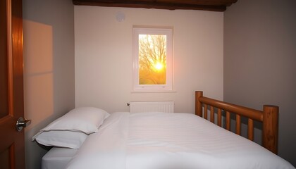 A small bedroom with a rustic wooden bed frame and clean white linens, with the soft golden light of an autumn sunset streaming in through the window.	