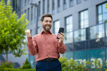 Cheerful mature businessman celebrating using smartphone outdoors. Man in professional outfit...
