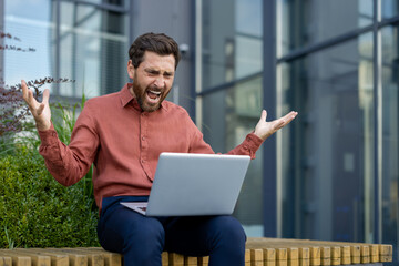 Mature businessman shows frustration and stress while working on laptop outdoors. Casual attire,...