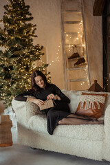 Obraz premium Young beautiful brunette woman in black dress lies on sofa with christmas gift near christmas tree