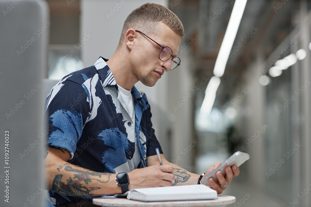 Poster Side view of young male business worker checking message on mobile phone writing in notebook while sitting at table in common area of modern office building, copy space