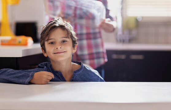 A young boy with brown hair smiles while leaning on a kitchen counter. The scene shows a cozy kitchen, with a blurred adult in the background preparing breakfast at dawn.