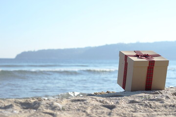 A beautifully wrapped gift box with a red plaid ribbon resting on a sandy beach, with distant...