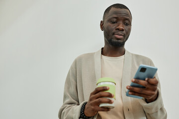 Medium shot of African American businessman in beige sweater outfit for office checking messages on phone captured with reusable coffee cup during break, copy space