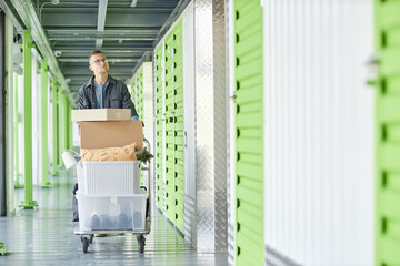 Full length shot of man pushing cart loaded with boxes full of home items walking along warehouse hallway in search of personal storage unit while moving out from apartment, copy space