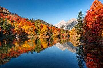 Obraz premium Tranquil lake surrounded by autumn trees and mountains, with vibrant red, orange, and yellow leaves mirrored on the water's surface, ai.