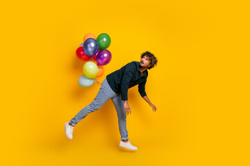 Playful young man in denim and black shirt floating with colorful balloons against bright yellow backdrop.