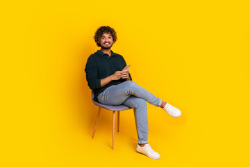Young man with curly hair sitting on chair against vibrant yellow background, smiling and looking at phone, wearing casual clothing