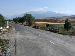 View of landcape with mountains and hills. Village near the mountains. Road among the trees leads into the mountain gorge. Beautiful clouds above mountains. Vivid natural scenery photo.