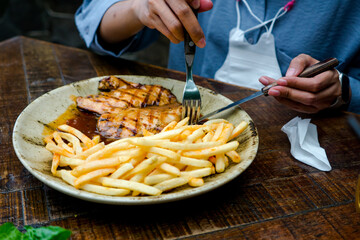 A woman enjoys a delicious chicken steak and French fries at a restaurant, served on a beautiful cream-colored plate with exquisite texture. The meal is placed on a stylish wooden table