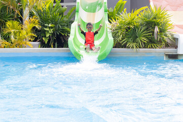 Asian little boy wearing a swimming suit and swimming goggles playing water slide at water park having fun on summer vacation.