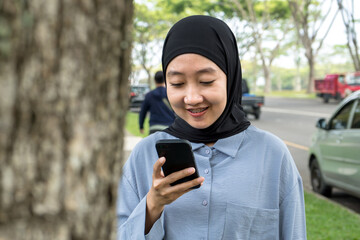 A joyful woman in a black hijab and blue shirt smiles brightly while holding a smartphone, standing at the edge of a beautiful park sidewalk