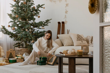 Stylish arrangement of a living room with Christmas decorations. A young woman wraps gifts by the...