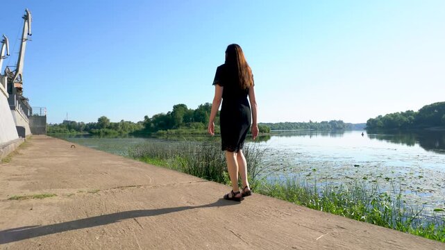 A woman in a black dress walks along the river embankment with her back to the camera.
