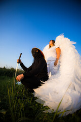 Naklejka premium Handsome samurai and angel against blue sky. A masked samurai kneels on a grassy field holding a katana next to a man with large white wings resembling an angel under a clear blue sky.