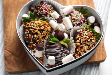Heart-shaped gift box containing chocolate-dipped strawberries topped with nuts, sprinkles and marshmallows on eco-friendly wooden boards. Perfect for celebrations. Top view