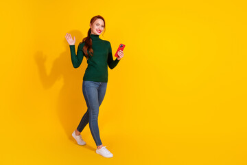 Full length photo of charming positive woman wear green turtleneck waving hi texting modern gadget empty space isolated yellow color background