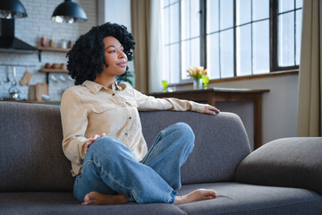 Young calm black woman relaxing sit on comfortable sofa in modern living room, lazy happy african...