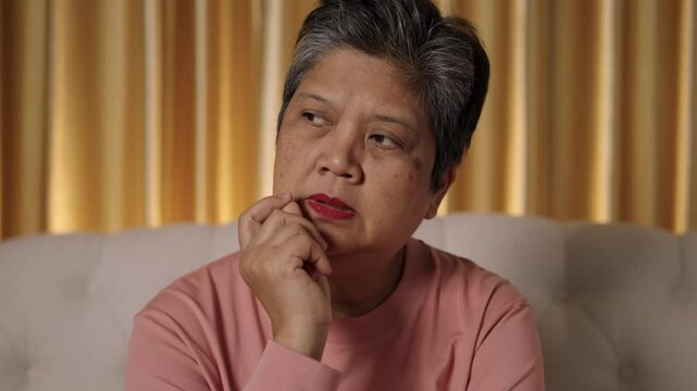 Close-up frontal video captures the face of an elderly Asian woman with short hair, hands resting on her cheeks and temples, who appears to be thinking while sitting on a sofa in a living room.